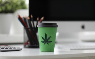 In an office on a desk there is a coffee to-go cup that is green and has a marijuana leaf decoration